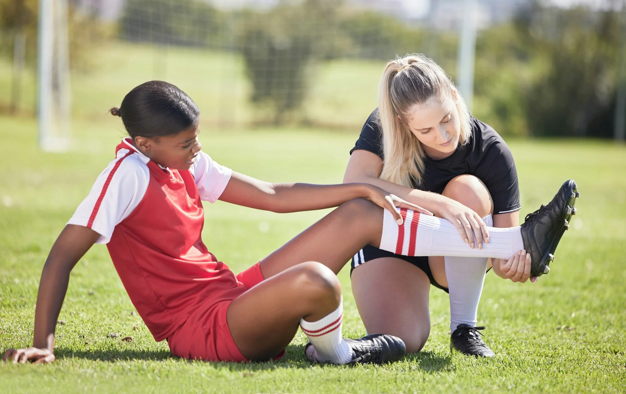 Female soccer player seated on field with teammate examining injured ankle